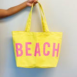Yellow tote bag with 'BEACH' written in pink on a light gray background