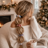 A woman with long blonde hair styled in a braid wearing colorful patterned hair ties, sitting in a cozy armchair by a fireplace with a Christmas tree in the background.