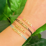 A slim satin thread bracelet with the word 'MAMA' written in golden letters, displayed on a person's wrist.