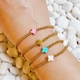 Gold bracelets with colorful star-shaped charms on a wrist, against a background of pebbles.