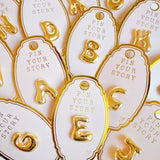 Glossy, balloon-style metal initial brooch pins in a playful, rounded design, available in multiple letters with a puffy, dimensional shape and metallic finish.