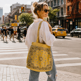 A woman on a city crosswalk carries a large yellow tote bag with a black paisley bandana print. She is wearing a white button-down shirt and light blue jeans, with a busy city street, yellow cabs, and pedestrians in the background.