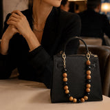 Black handbag with wooden bead handle held by a person in a blurred indoor setting