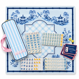 A Mahjong set arranged on a decorative blue-and-white porcelain-style mat, with a striped carrying case, tiles, dice, and a stack of score cards. The case has pink handles and the word "MAHJ" visible on the front.