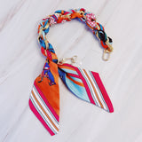 Stylish braided scarf lanyard featuring a gold-tone chain intertwined with patterned fabric, designed to securely attach to a phone while doubling as a chic fashion accessory.