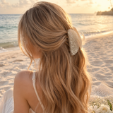 Woman with long blonde hair tied back with a decorative hairpiece on a beach at sunset.