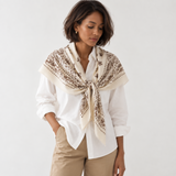 A woman wearing a cream and brown paisley patterned silk scarf tied around her neck over a white button-down shirt and beige trousers, photographed against a clean white studio background.