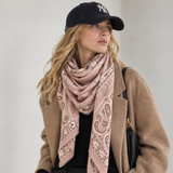 A woman wearing a dusty pink paisley bandana scarf styled with a camel coat, black pants, a black baseball cap, and a black shoulder bag against a neutral light gray background.