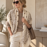 A woman wearing a cream shawl with a brown paisley pattern over a taupe sweater and white pants, styled in a warm, modern interior setting.