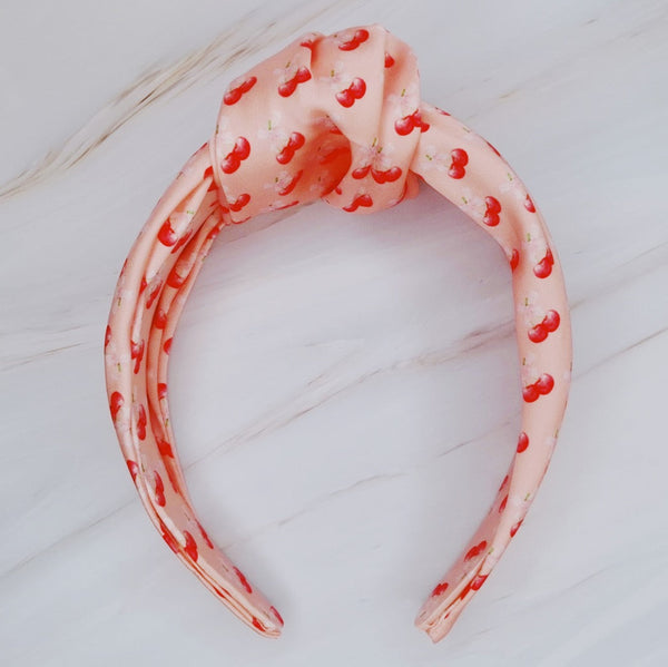 Soft satin headband adorned with a playful cherry and bow pattern, featuring vibrant red cherries and delicate bows on a pink background.