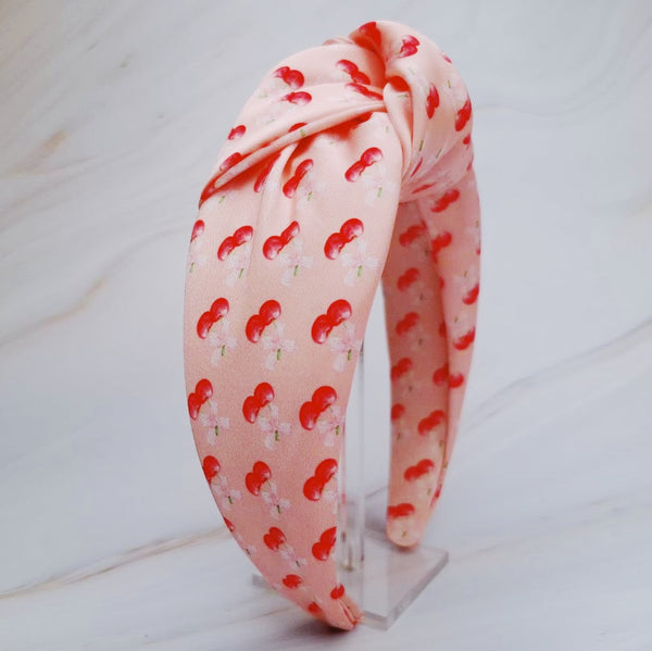 Soft satin headband adorned with a playful cherry and bow pattern, featuring vibrant red cherries and delicate bows on a pink background.