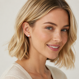 A close-up portrait of a smiling blonde woman wearing a small round stud earring on her left ear, against a soft light gray background.
