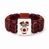 A beaded bracelet featuring glossy, rounded rectangular beads in shades of deep red, burgundy, and terracotta, arranged in a circle around a central square white tile. The tile is decorated with a red floral vase motif with black leaves.