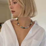 A woman with a short blonde bob wearing a multi-colored beaded necklace and a white button-down shirt, shown in a close-up profile view.
