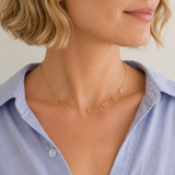 A close-up of a woman wearing a delicate gold necklace with the name 'COOL MOM' spelled out in small gold letters, paired with a light blue button-down shirt.