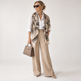 A woman models a stylish outfit featuring a beige and black floral patterned scarf draped over her shoulders, paired with a white button-down shirt, beige wide-leg trousers, and a taupe handbag against a plain light gray studio background.