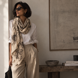 A woman wearing a cream and black botanical print scarf, a white button-down shirt, and beige trousers stands in a sunlit room next to a window.