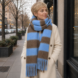 A woman standing on a city sidewalk wearing a cream coat and a thick knit scarf with wide brown and light blue stripes.