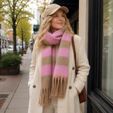 A woman standing on a city sidewalk wearing a cream coat, a beige baseball cap, and a thick knit scarf with wide pink and beige stripes and fringe ends.
