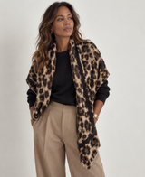 A woman modeling a leopard print wrap jacket over a black long-sleeve top and beige wide-leg trousers, photographed against a plain light gray studio background.