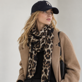 A woman wearing a beige camel coat, black top and trousers, a leopard print scarf, a black baseball cap, and carrying a black shoulder bag against a light gray studio background.