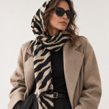 A woman wearing a beige and black zebra-print scarf, a camel-colored coat, black trousers, and black sunglasses, photographed against a plain light background.