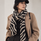 A woman wearing a black baseball cap, a black and beige zebra-patterned scarf, a camel coat, and black trousers, carrying a black shoulder bag against a plain light background.