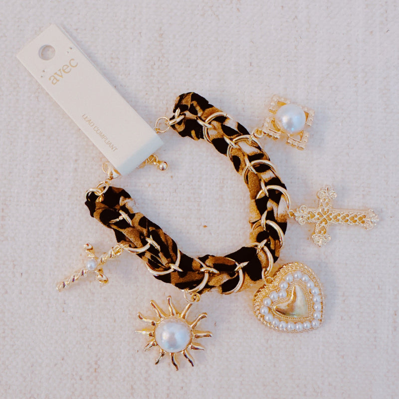 Gold-tone charm bracelet with a leopard-print scarf wrap, featuring dangling heart, cross, sun, and pearl-accented charms.