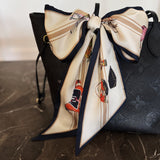 Black handbag with a decorative bow featuring cartoon characters on a marble surface. Silky jumbo twilly scarf featuring an equestrian-inspired print with horses and carriage motifs, finished with a contrasting navy border for styling around the neck or handbag.