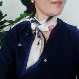 Person wearing a patterned scarf with a plant in the background. Silky twilly scarf featuring an equestrian-inspired print with horses and carriage motifs, finished with a contrasting navy border for styling around the neck or handbag.