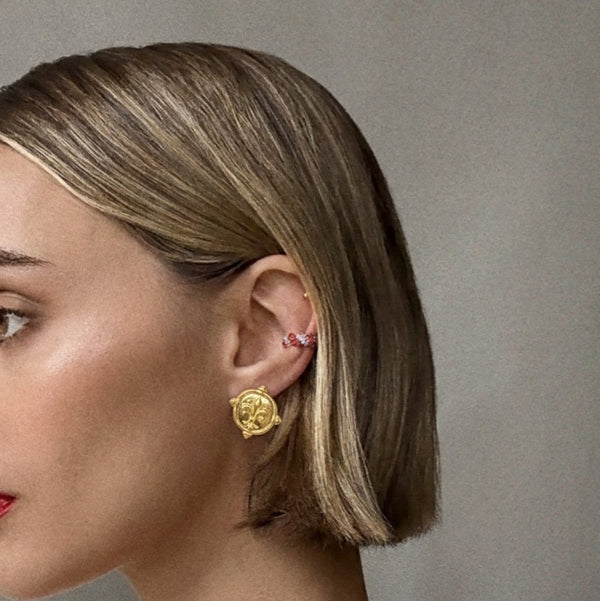 Gold round stud earrings featuring a raised fleur-de-lis design on a coin-style medallion with decorative beaded edges.