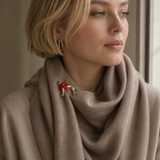 A close-up of a woman wearing a taupe knit cowl scarf with a gold dog-shaped brooch featuring a red heart on its chest, styled with gold hoop earrings.