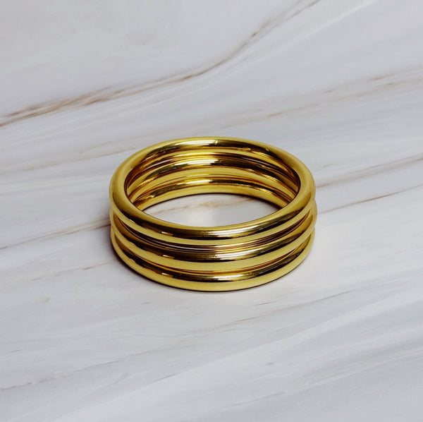  Set of five shiny gold-tone bangles stacked together on a neutral marble background, showcasing a sleek and polished design.