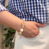 Person wearing a bracelet with a cross and pearls, standing in front of a plant.