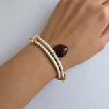 A close-up of a person's wrist wearing a double-strand braided cream rope bracelet with gold-toned hardware and a small dark brown heart-shaped stone charm.