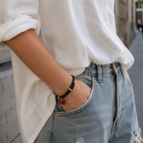A person wearing a white linen button-up shirt and light-wash distressed jeans, with one hand in a pocket and the other showing a black bracelet with gold hardware and a small red heart charm.