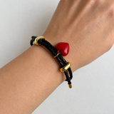 A close-up of a person's wrist wearing a black braided cord bracelet with gold-toned hardware and a small glossy red heart charm, against a plain light background.