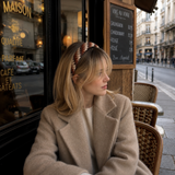 A woman with blonde hair sits at a cafe table wearing a beige coat and a brown and white patterned headband, with a cafe window and street scene in the background.