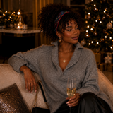 A woman sits on a couch in a cozy, warmly lit living room, wearing a gray knit button-up sweater and black pants, holding a glass of champagne. She has a plaid headband and a small black handbag with a gold chain.