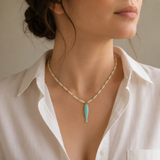 A close-up of a woman wearing a delicate necklace with a turquoise fish pendant and cream-colored oval beads on a white collared shirt.