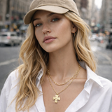Woman wearing a beige cap and white shirt with gold jewelry on a city street. Layered necklace featuring two delicate chains with a polished cross pendant, available in multiple metal tone variations for a modern, faith-inspired look.