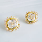 Elegant stud earrings featuring a stone encircled by small sparkling crystals, all set in a polished gold frame. The cushion-cut center gem catches the light beautifully, creating a timeless and sophisticated look.
