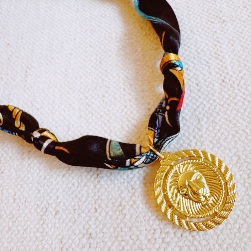 Elegant scarf necklace featuring a bold medallion pendant and luxe printed fabric intertwined with a chain, combining statement style with wearable versatility.