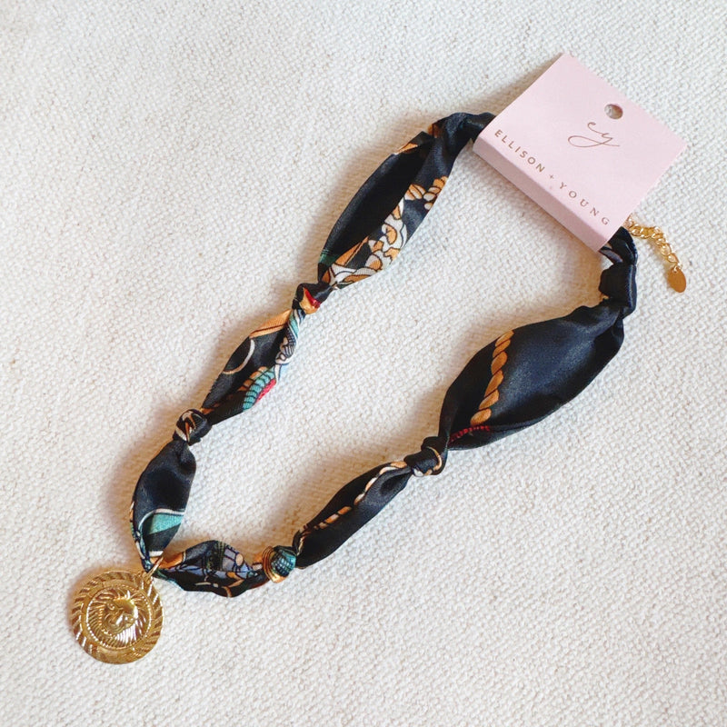 Elegant scarf necklace featuring a bold medallion pendant and luxe printed fabric intertwined with a chain, combining statement style with wearable versatility.