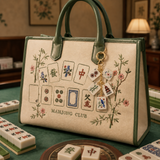 A structured beige handbag with dark green handles and trim, decorated with embroidered mahjong tiles and floral motifs, resting on a green mahjong table with tiles visible around it.