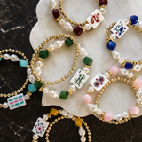 A styled flat-lay of multiple beaded bracelets arranged on a white scalloped tray over a dark marble surface. The bracelets feature gold-tone bead strands with white pearl-like beads, green and pink faceted beads, and white square tiles printed with colorful Chinese mahjong-style symbols and characters.