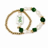 A beaded stretch bracelet featuring gold-tone ball beads, white baroque pearls, faceted dark green beads, and a central white tile charm with green bamboo graphics, displayed on a clean white background.