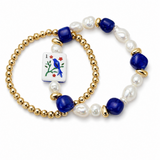 A double-strand beaded bracelet set on a white background. One bracelet features gold-tone ball beads with dark blue and white baroque pearls, and a small square card charm depicting a blue bird on a red suit of cards.