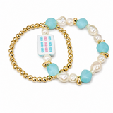 A pair of gold beaded stretch bracelets with light blue faux-pearl beads, white freshwater pearl beads, and a small white tile charm featuring colorful mahjong-style symbols, displayed on a clean white background.