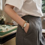 A person wearing a white short-sleeve button-up shirt and gray trousers, with one hand in their pocket, showcasing a double-strand beaded bracelet. The bracelet features gold-toned round beads, white beads with green accents, and a white tile charm depicting a green and white mahjong tile.